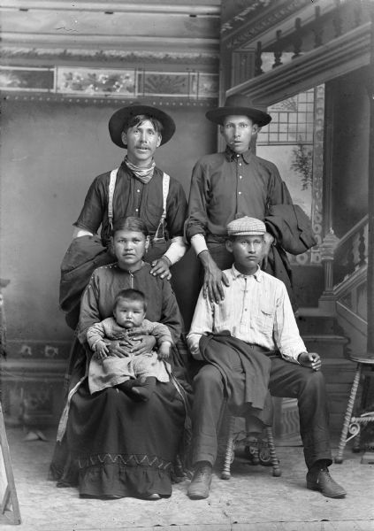 A studio portrait of a group of Native Americans. A woman in a dress holds an infant in her lap, a man stands behind her, and two young men, one standing and one seated next to them. All the men wear hats, and are holding their suit jackets. One man is smoking. In the background is a painted backdrop. Standing (l to r), Amos Wallace (WeeKah) and William Rainbow. Sitting, left to right, Belle (Youngswan) Standingwater, Whitepigeon, Wallace (WaNekMonkEKayWinKah), her baby, Tom Johnson Wallace (WaukJaHeSepKah), and Thomas Rainbow.