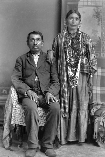 A studio portrait of an elderly Native American couple, with the husband seated wearing a suit jacket and trousers, and his wife standing to his left in front of a painted backdrop. She is wearing a dress with earrings and a necklace.