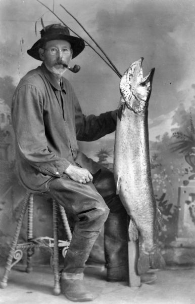 A studio portrait of an elderly man seated with a pipe in his mouth holding a large fish (probably a Muskellunge or Northern Pike) in front of a painted backdrop.