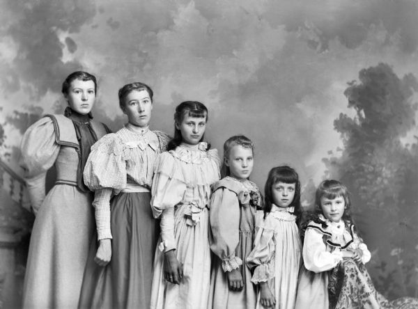 A studio portrait of six girls arranged in decreasing height, standing in front of a painted backdrop.