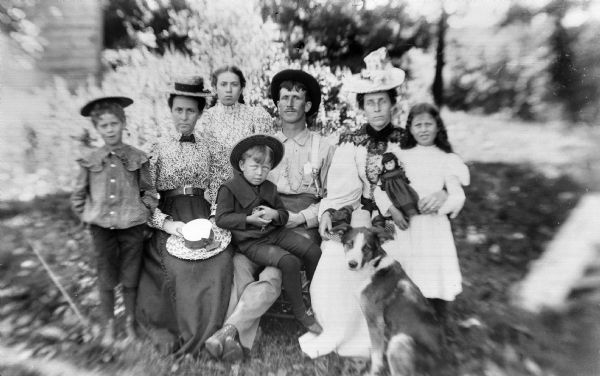 An outdoor family portrait with two women, one man, and four children. Two boys are wearing hats, and one of the girls is holding a doll. A dog is sitting in front of the family.