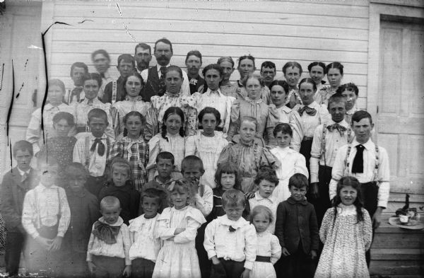 An outdoor portrait of a large group of men, women, and children posing in front of the wall of a frame building.
