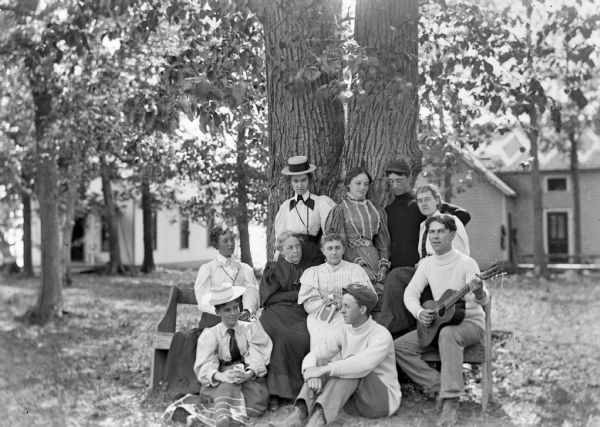A group of seven women and three men, some standing, and some sitting on a bench and on the ground, gathered under a tree in front of two frame buildings. One man is playing a guitar.