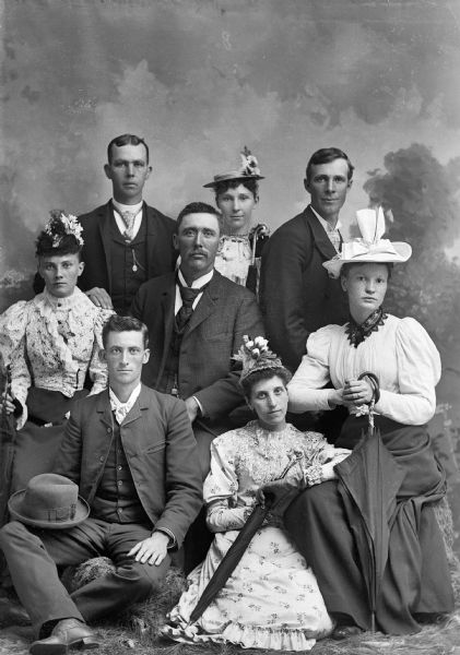 Studio portrait of four men and four women posing in front of a painted backdrop. All of the women are wearing hats and holding umbrellas. The men are wearing suit jackets, vests, neckties, and trousers.