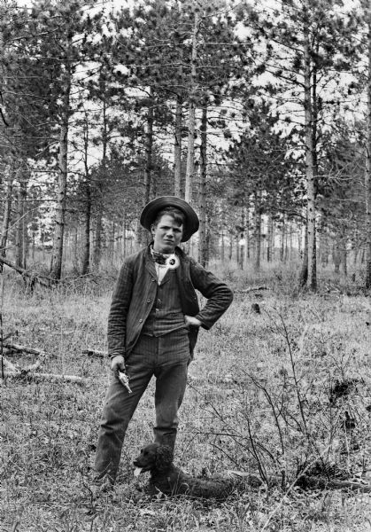 A portrait of a boy in the woods wearing a hat, jacket, and vest, holding a pistol in his right hand. There is a dog lying at his feet.