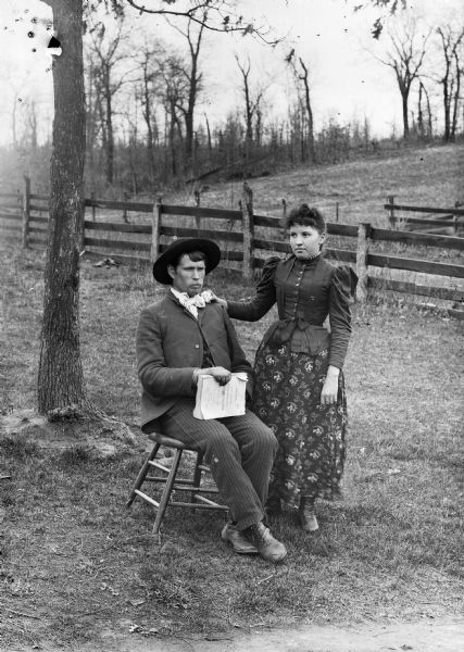An outdoor portrait of a man wearing a hat, suit jacket, and trousers seated on a stool, and a woman standing next to him, with a fence behind them. The man, presumably a violinist, is holding a book titled "J.W. Pepper's Universal Dancing Master, Prompter's Call-Book Violinists Guide."