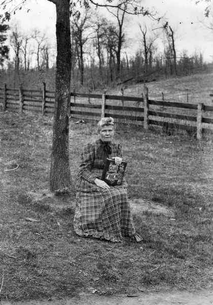 An outdoor portrait of a woman seated near a tree, holding a book titled "Our Friends," with a fence in the background.