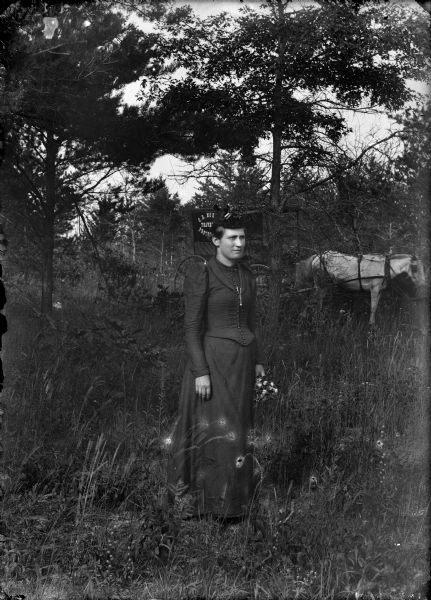 A portrait of a young woman wearing a hat and dress, and holding a bouquet of flowers, standing in a clearing. In the background is the darkroom horse-drawn wagon of C.R. Monroe, Traveling Photographer.