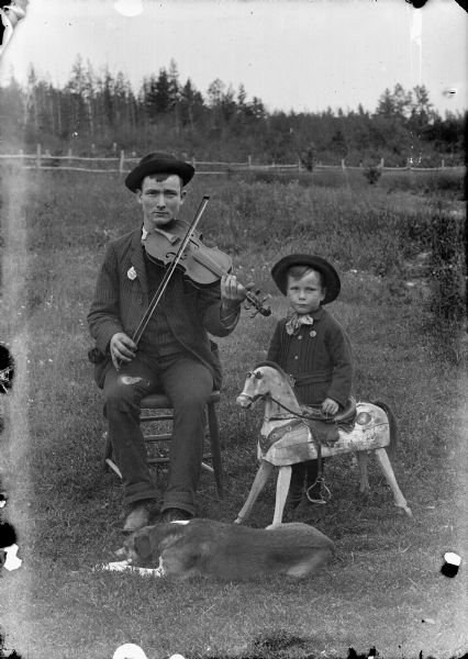 An outdoor portrait of a seated man holding a violin and bow. A small boy with a toy wooden horse stands next to him. A dog is resting in the grass in front of them.