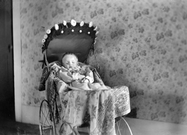 A portrait of a baby lying in a baby carriage decorated with flowers, possibly deceased.