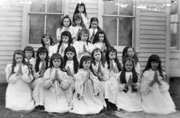 A portrait of 18 young girls wearing white dresses posing with their hands folded while kneeling in front of a building.