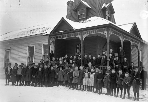A large group of men, women, and children of varying ages standing around the porch of a frame house in winter.