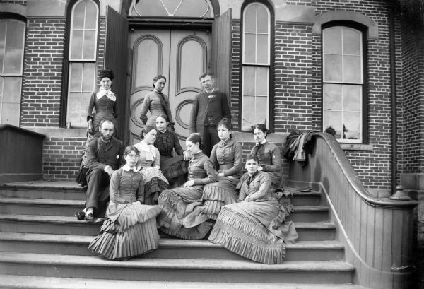 A group of two men and nine women posing on the steps leading into a brick building.