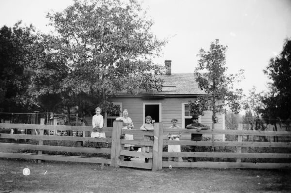 A portrait of a family, a father, mother, and three daughters, standing in their yard behind a wooden fence in front of a small house.