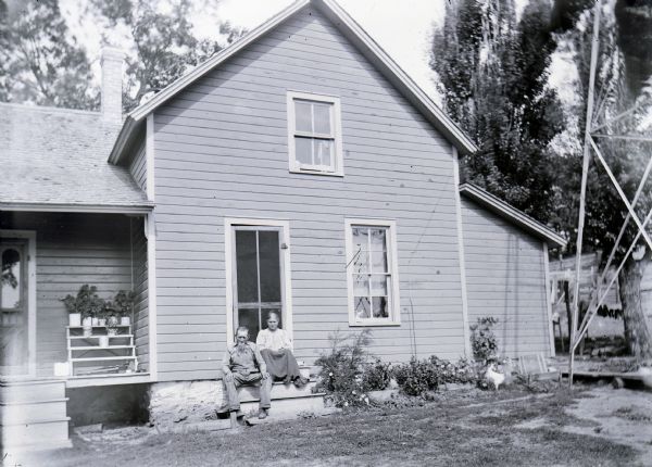 A portrait of an elderly couple sitting on steps leading into a frame house. On the right is a metal structure that is probably part of a windmill.