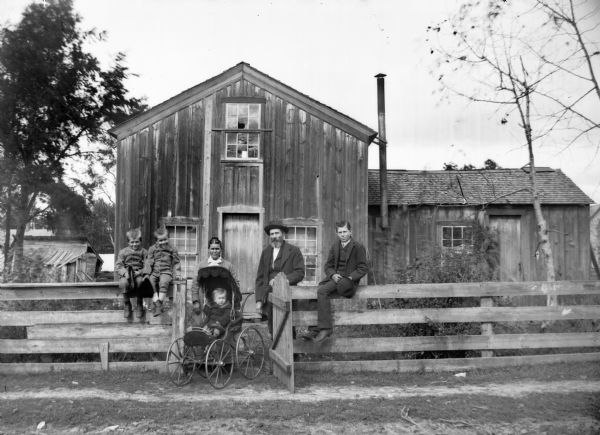 Three boys sitting on a wooden fence near a gate where a women and man are standing next to a baby carriage with a seated infant, in front of a frame house and other farm buildings.