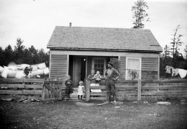 A portrait of a family of five, husband, wife, two daughters and an infant, standing by a fence in front of a small house. Laundry is hanging on a clothesline on the left.