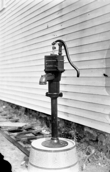 Still-life of a hand-pump, manufactured by Barber and Colman, Rockford, Illinois, attached to the top of a barrel near the side of a building.