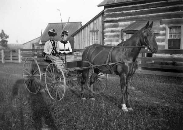 A portrait of two women sitting in a horse-drawn buggy stopped in front of a wooden fence and a log house. Photographer's shawdow is in foreground.