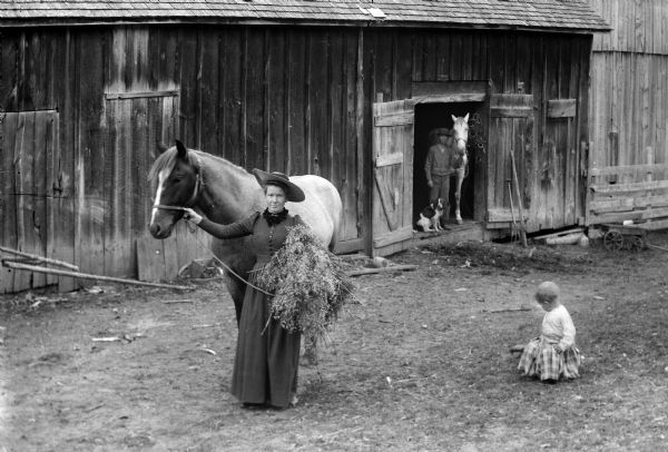 A portrait of a woman in a straw hat holding a horse in a barnyard and carrying hay in her left hand. There is an infant on the ground to her left, and a boy standing in the doorway behind her posing with a horse with a dog.