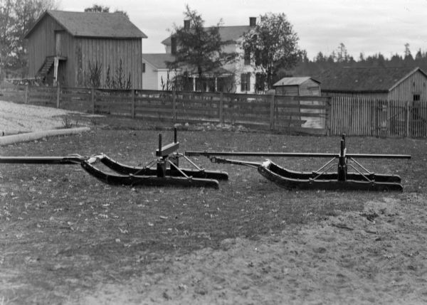 An empty bobsled in a farmyard in front of a wooden fence, a frame house, and other farm buildings.
