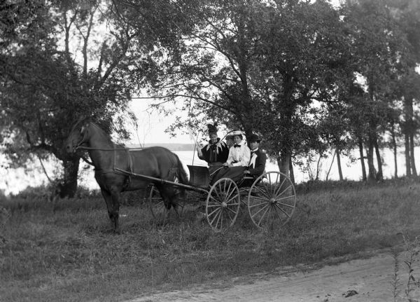 Group seated in a horse-drawn buggy are on the side of a road next to a river. The two women are sitting on the laps of the two men.