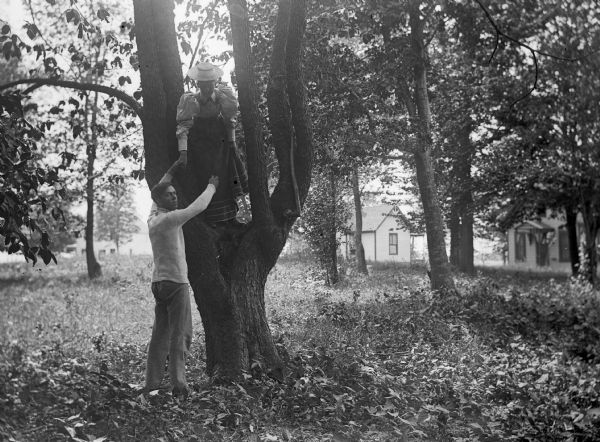 A man is helping a young woman down from a tree in a clearing. There are cabins or small houses in the background.