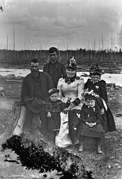 A family of six posing outdoors on the shoreline of a river with a cleared forest in the background. The husband is sitting on a tree stump, and the wife two girls are sitting on boxes. A boy and girl stand behind them.