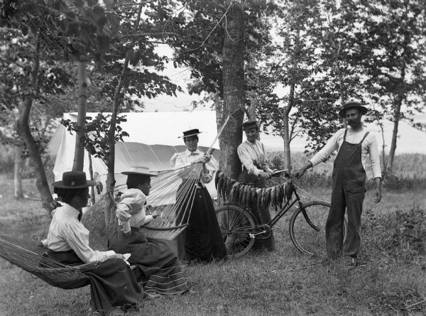 A group of four women and one man gathered around a hammock and bicycle. The man is displaying a stringer of fish which is attached to the hammock. There is a large tent in the background.