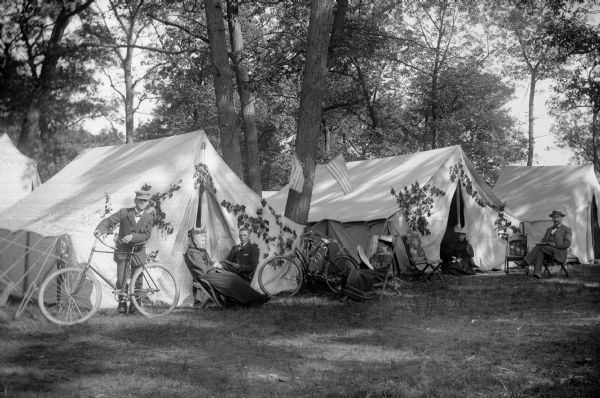 A group of three women and three men relaxing near tents decorated with foliage and American flags. One of the men is holding a bicycle.