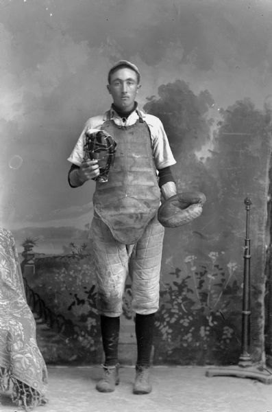 A studio portrait of a man wearing a baseball catcher's padded uniform and catchers glove, holding a face mask in his right hand. He is standing in front of a painted backdrop.