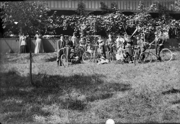 Group posed standing and sitting on grass with bicycles and oars in front of an open wooden building with covered hanging vines. One man is holding a set of oars.