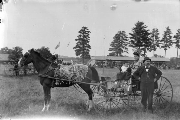 Woman and child posed sitting in a buggy pulled by a single horse wearing a fly-net. A man is standing in front of them. The grandstand at the Jackson County Fairgrounds is in the background.