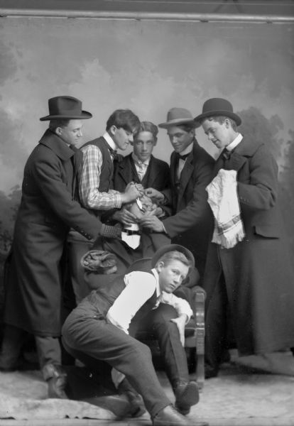 Studio portrait of six young men pretending to pull a seated young man's tooth with pliers. One man is on the floor holding the dental patient's legs. Three men are in overcoats and four are wearing hats. There is a painted backdrop behind them.