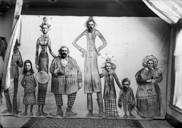 A group portrait in a studio of four women and three men posing behind a canvas with comical drawings of characters with the faces cutout.