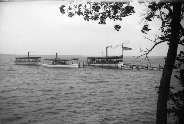 View from shoreline of three steam-powered passenger boats approaching a dock with a flag on it.