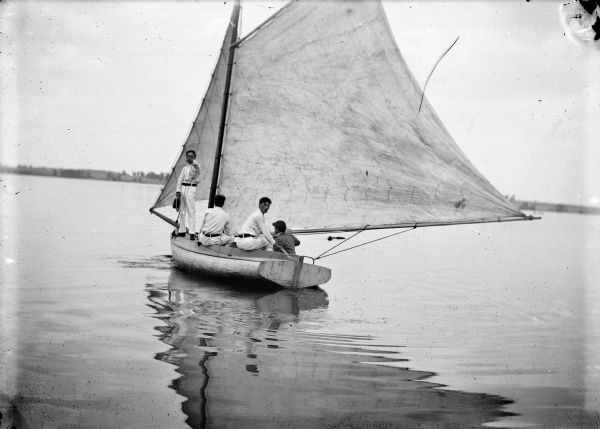 Four men sailing a small boat on a lake, with a shoreline in the background.
