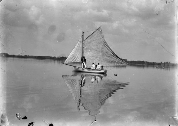 View from water of five men are sailing a small boat on a lake. There is another smaller boat on the right, and a shoreline in the background.