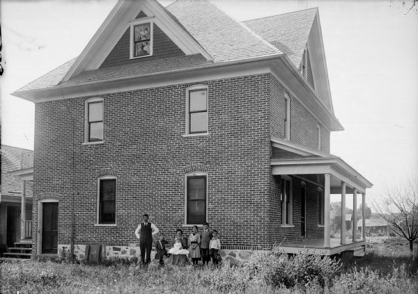 A family is posed outside their three-story brick home in the yard. A woman in the middle sits in a chair with a child in her lap. Four other children stand beside her, and a man with a moustache stands on the left.