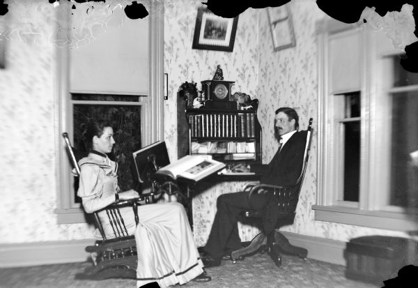 Portrait of a man and a woman posed sitting in the corner of a room near a desk and two book stands. The desk has a clock and many books on it.