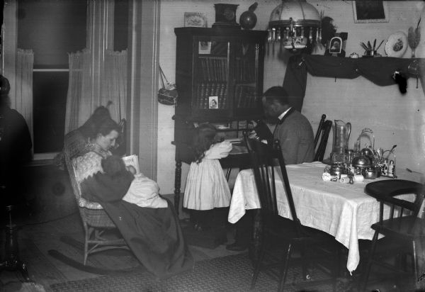 A family is posed in a dining room with a woman seated and holding a small child. A man is seated at a desk in the corner, with a young girl standing nearby. There is also another seated person on the extreme left side.