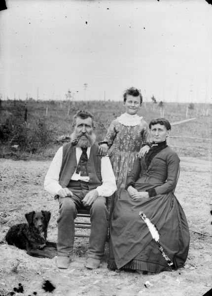 Outdoor portrait of a man and woman posed sitting with a young girl standing between them. A dog is lying on the ground. The woman and girl are wearing dresses, and the man wears a necktie, vest, and trousers.