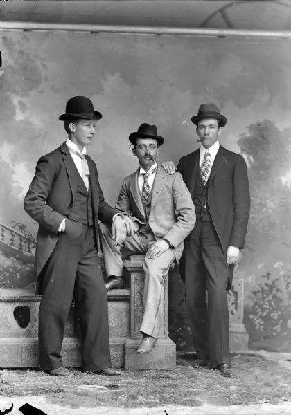 Studio portrait of a man sitting on a stone wall between two other standing men in front of a painted backdrop. They are wearing suit jackets, neckties, and hats, and have cigars.