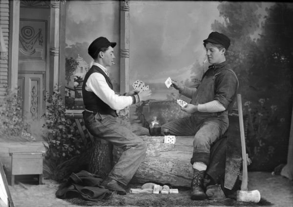 A studio portrait of two men, probably lumberjacks, that are posed playing cards, drinking, and smoking in front of a painted backdrop. The two men are sitting on a log with a saw and an axe nearby. The man on the left is wearing a vest, trousers, and hat, and the man on the right wears overalls and a hat.