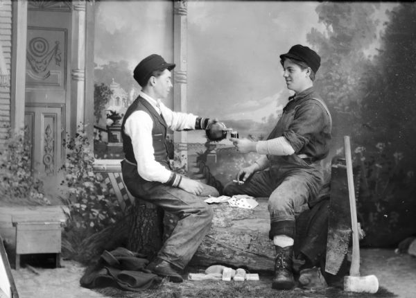 Two unidentified white men, dressed as lumberjacks, sit on a wood log in Van Schaick's studio, drinking, smoking and playing cards, with a saw and an axe nearby. The man on the left side is wearing a hat, vest, and trousers and pouring the other man wearing overalls a drink.