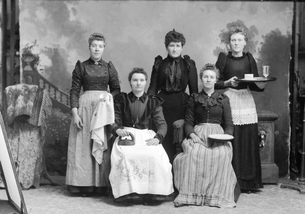 Studio portrait of two women sitting in front of three standing women, all holding household items, including: pie, iron, cup, towel, platter, and glasses. They are in front of a painted backdrop. All five women are wearing dresses, and four of them wear aprons.