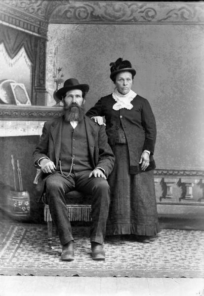A studio portrait of a seated elderly man with a beard, and an elderly woman in a dress standing beside him. Both are wearing hats in front of a painted backdrop. He is wearing a suit jacket, vest with a watch fob, and trousers.