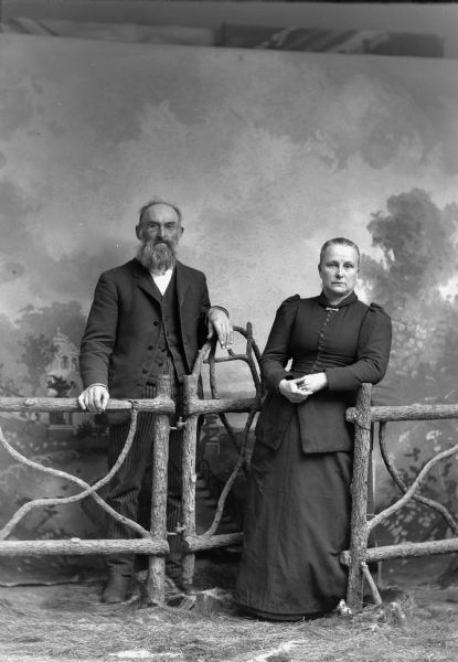 Studio portrait of a man with a beard, wearing eyeglasses, suit jacket, vest, and trousers, and a woman in a long dress. They are standing by a wooden fence in front of a painted backdrop.