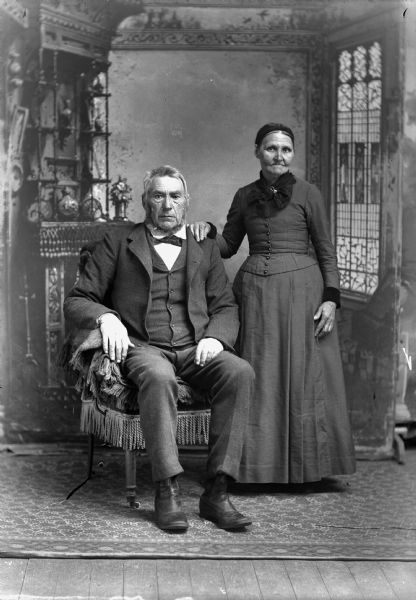 Studio portrait of a seated elderly man wearing a suit jacket, vest, necktie, and trousers, and an elderly woman in a long dress with a lace collar, standing beside him. They are in front of a painted backdrop.