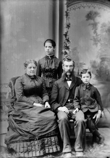 Studio portrait of R.B. Jones and family. The man has a long beard and moustache, and is wearing a suit jacket, vest with watch fob, necktie and trousers. Next to him is a young boy also wearing a suit jacket and necktie. The woman, seated, is wearing a dress. A girl in a dress stands behind them. They are in front of a painted backdrop.